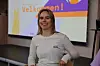 En smilende blond kvinne sitter foran en presentasjonsskjerm i et klasserom.
