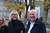 To smilende voksne står utendørs foran trær og universitetsbygninger en høstdag.
